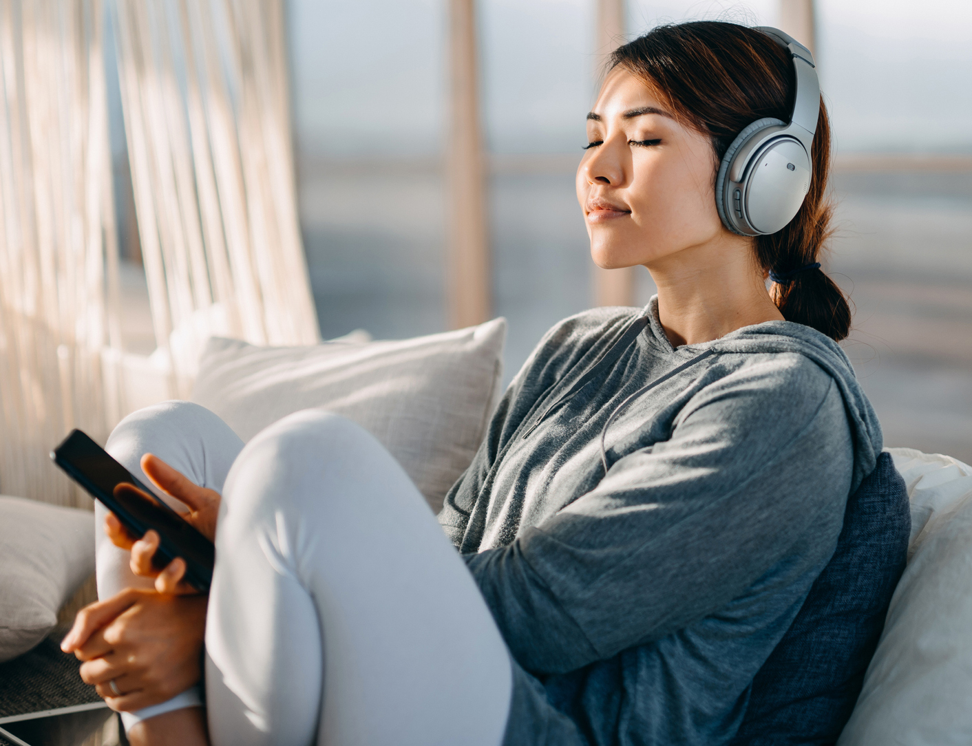 Relaxed young Asian woman with eyes closed sitting on her bed enjoying music over headphones from smartphone at home Capsulectomy in Bucks County, PA & Hunterdon County, NJ